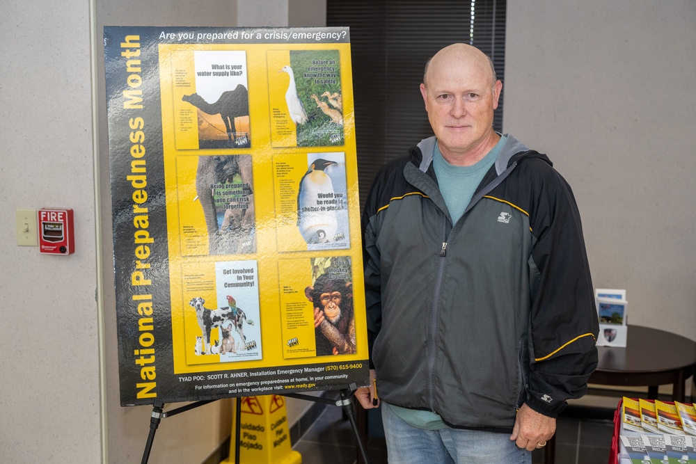 A middle-aged bald man stands beside a yellow and black 'National Preparedness Month' display board in what appears to be an indoor facility. The poster features animal imagery and emergency preparedness messaging, asking 'Are you prepared for a crisis/emergency?' A fire alarm is visible on the wall behind him, and informational brochures are displayed on a table nearby.