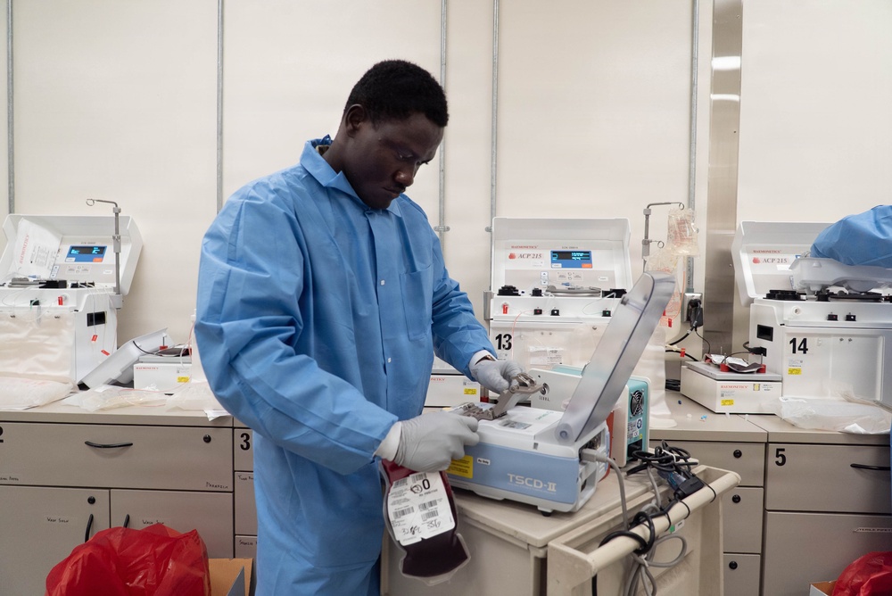 A laboratory technician wearing a blue lab coat and white gloves operates a TSCD-II device, likely a sterile tubing welder used in blood processing. The setting appears to be a blood bank or transfusion medicine laboratory, with multiple ACP 215 automated cell processing machines visible in the background. A blood bag labeled with type 'O' is visible in the technician's hand.