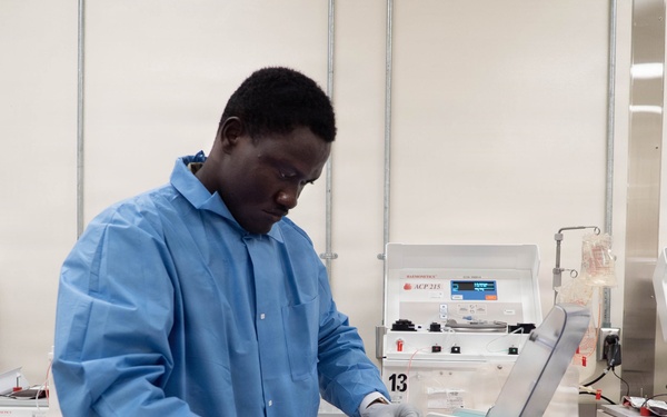 A laboratory technician wearing a blue lab coat and white gloves operates a TSCD-II device, likely a sterile tubing welder used in blood processing. The setting appears to be a blood bank or transfusion medicine laboratory, with multiple ACP 215 automated cell processing machines visible in the background. A blood bag labeled with type 'O' is visible in the technician's hand.