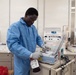 A laboratory technician wearing a blue lab coat and white gloves operates a TSCD-II device, likely a sterile tubing welder used in blood processing. The setting appears to be a blood bank or transfusion medicine laboratory, with multiple ACP 215 automated cell processing machines visible in the background. A blood bag labeled with type 'O' is visible in the technician's hand.