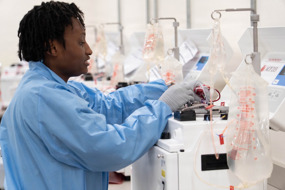 A laboratory technician wearing a blue lab coat and gray gloves is operating medical blood processing equipment in what appears to be a blood bank or transfusion medicine facility. Multiple automated cell processing machines are visible in a row, each equipped with hanging IV-style blood collection bags containing red fluid. The setting suggests a high-throughput blood processing or apheresis operation.