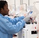 A laboratory technician wearing a blue lab coat and gray gloves is operating medical blood processing equipment in what appears to be a blood bank or transfusion medicine facility. Multiple automated cell processing machines are visible in a row, each equipped with hanging IV-style blood collection bags containing red fluid. The setting suggests a high-throughput blood processing or apheresis operation.