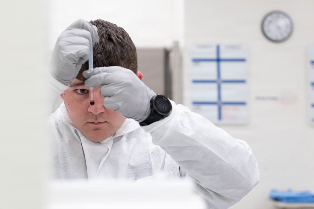 A laboratory technician or scientist wearing a white cleanroom suit and gray nitrile gloves is closely examining a small vial or test tube held up near eye level. The individual appears focused and is wearing a black digital watch. The setting is a clean, well-lit laboratory environment with administrative charts and a wall clock visible in the background.