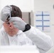 A laboratory technician or scientist wearing a white cleanroom suit and gray nitrile gloves is closely examining a small vial or test tube held up near eye level. The individual appears focused and is wearing a black digital watch. The setting is a clean, well-lit laboratory environment with administrative charts and a wall clock visible in the background.