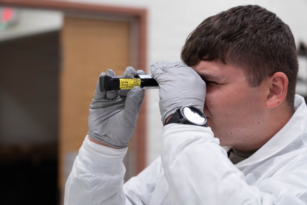 A young male technician wearing a white protective suit and gray nitrile gloves is closely examining a small yellow-labeled vial or tube by holding it up to his eye. The item appears to have printed text and labeling visible on its yellow surface. The setting appears to be an indoor facility with a blurred background.