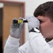 A young male technician wearing a white protective suit and gray nitrile gloves is closely examining a small yellow-labeled vial or tube by holding it up to his eye. The item appears to have printed text and labeling visible on its yellow surface. The setting appears to be an indoor facility with a blurred background.