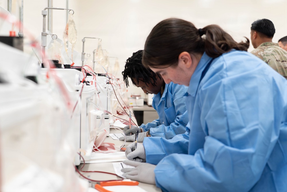 The image shows multiple individuals in blue laboratory gowns and gray gloves working at a long bench equipped with blood collection or processing equipment. They appear to be engaged in a medical or blood banking procedure, with IV bags, tubing, and collection devices visible. In the background, individuals in military camouflage uniforms are also present, suggesting a military medical training or operational setting.