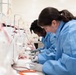 The image shows multiple individuals in blue laboratory gowns and gray gloves working at a long bench equipped with blood collection or processing equipment. They appear to be engaged in a medical or blood banking procedure, with IV bags, tubing, and collection devices visible. In the background, individuals in military camouflage uniforms are also present, suggesting a military medical training or operational setting.