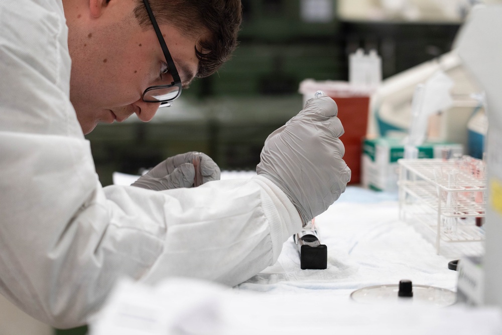 A young male laboratory technician or scientist is performing a precise procedure at a lab bench, carefully manipulating what appears to be a small instrument or sample holder. He is wearing standard laboratory personal protective equipment including a white lab coat and latex gloves, and is focused intently on his work. The background reveals a well-equipped laboratory setting with test tubes, reagent bottles, and other lab supplies.