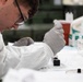 A young male laboratory technician or scientist is performing a precise procedure at a lab bench, carefully manipulating what appears to be a small instrument or sample holder. He is wearing standard laboratory personal protective equipment including a white lab coat and latex gloves, and is focused intently on his work. The background reveals a well-equipped laboratory setting with test tubes, reagent bottles, and other lab supplies.