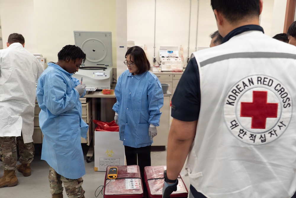 The image shows a medical laboratory setting where personnel from the Korean Red Cross and military/medical staff are collaborating. A person wearing a Korean Red Cross vest is observing alongside individuals in blue medical gowns and military camouflage pants. The scene appears to involve blood or biological sample handling, with biohazard-labeled containers and medical transport cases visible.