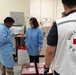 The image shows a medical laboratory setting where personnel from the Korean Red Cross and military/medical staff are collaborating. A person wearing a Korean Red Cross vest is observing alongside individuals in blue medical gowns and military camouflage pants. The scene appears to involve blood or biological sample handling, with biohazard-labeled containers and medical transport cases visible.