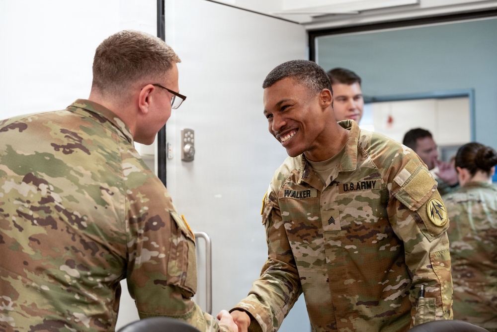Two U.S. Army soldiers in OCP (Operational Camouflage Pattern) uniforms are shaking hands in what appears to be an indoor military facility. The soldier on the right, identified by his name tape as 'Walker' with the rank of Sergeant, is smiling broadly during the handshake. Several other military personnel are visible in the background.
