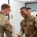 Two U.S. Army soldiers in OCP (Operational Camouflage Pattern) uniforms are shaking hands in what appears to be an indoor military facility. The soldier on the right, identified by his name tape as 'Walker' with the rank of Sergeant, is smiling broadly during the handshake. Several other military personnel are visible in the background.
