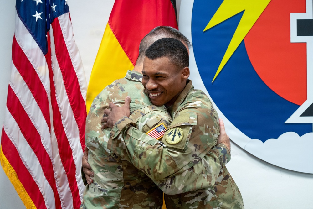 Two U.S. Army soldiers in OCP (Operational Camouflage Pattern) uniforms are embracing in what appears to be an official military ceremony or event. The soldier facing the camera is smiling broadly, suggesting a celebratory or farewell occasion. The setting includes national flags and a military unit insignia on the wall behind them.