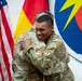 Two U.S. Army soldiers in OCP (Operational Camouflage Pattern) uniforms are embracing in what appears to be an official military ceremony or event. The soldier facing the camera is smiling broadly, suggesting a celebratory or farewell occasion. The setting includes national flags and a military unit insignia on the wall behind them.