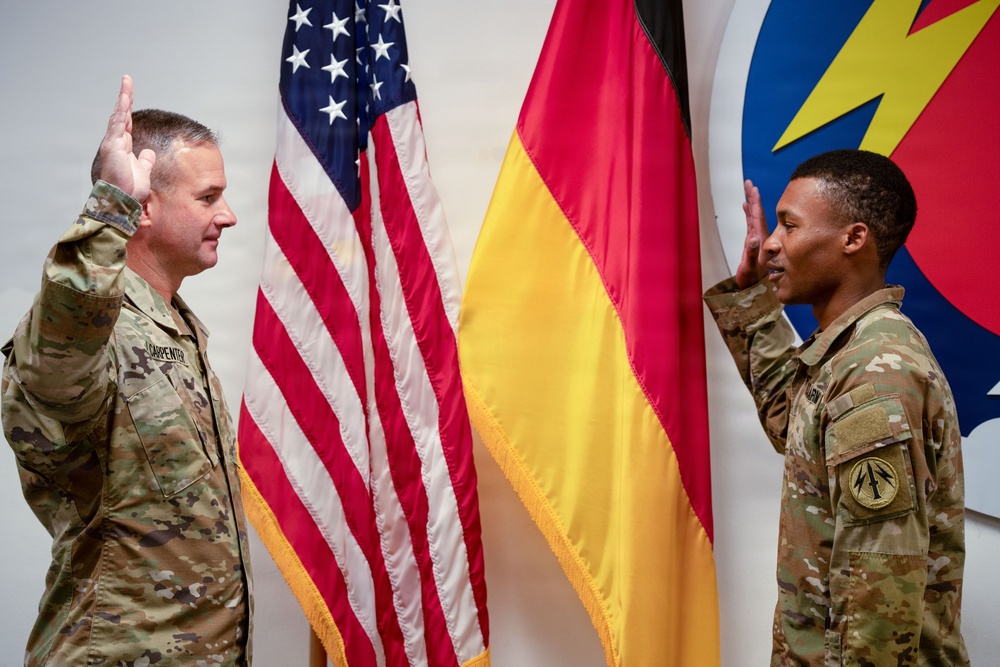 Two U.S. Army soldiers in MultiCam/OCP camouflage uniforms are conducting what appears to be a military oath or reenlistment ceremony, both with right hands raised. They are standing in front of an American flag and a German flag, with a military unit insignia (featuring a lightning bolt) visible on the wall behind them. The name tape 'CARPENTER' is visible on the left soldier's uniform.