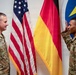 Two U.S. Army soldiers in MultiCam/OCP camouflage uniforms are conducting what appears to be a military oath or reenlistment ceremony, both with right hands raised. They are standing in front of an American flag and a German flag, with a military unit insignia (featuring a lightning bolt) visible on the wall behind them. The name tape 'CARPENTER' is visible on the left soldier's uniform.