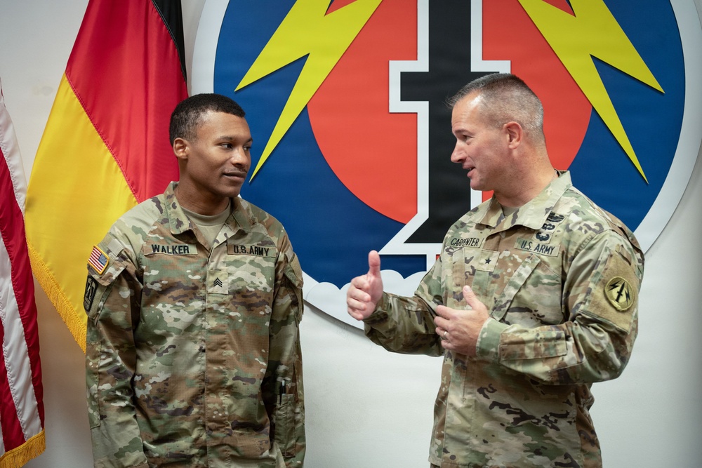 Two U.S. Army soldiers in OCP (Operational Camouflage Pattern) uniforms are engaged in conversation. The soldier on the left, identified by name tape as 'WALKER' with Sergeant rank insignia, is listening while the soldier on the right, identified as 'CARPENTER' with a Brigadier General star visible, gestures with a thumbs-up. They are standing in front of a German flag and what appears to be a 56th Signal Battalion or similar unit insignia backdrop.