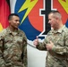 Two U.S. Army soldiers in OCP (Operational Camouflage Pattern) uniforms are engaged in conversation. The soldier on the left, identified by name tape as 'WALKER' with Sergeant rank insignia, is listening while the soldier on the right, identified as 'CARPENTER' with a Brigadier General star visible, gestures with a thumbs-up. They are standing in front of a German flag and what appears to be a 56th Signal Battalion or similar unit insignia backdrop.