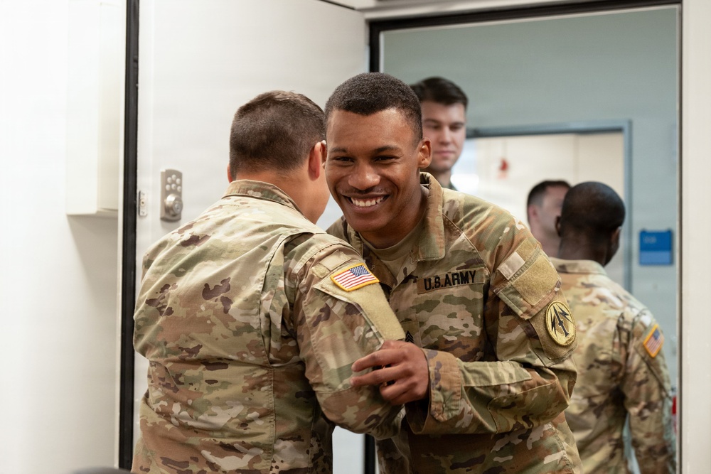 Two U.S. Army soldiers in MultiCam (OCP) uniforms are engaged in a friendly interaction, with one soldier smiling broadly while the other appears to be pinning or adjusting something on his sleeve. Several other soldiers are visible in the background near a doorway. The setting appears to be an indoor military facility.