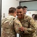 Two U.S. Army soldiers in MultiCam (OCP) uniforms are engaged in a friendly interaction, with one soldier smiling broadly while the other appears to be pinning or adjusting something on his sleeve. Several other soldiers are visible in the background near a doorway. The setting appears to be an indoor military facility.