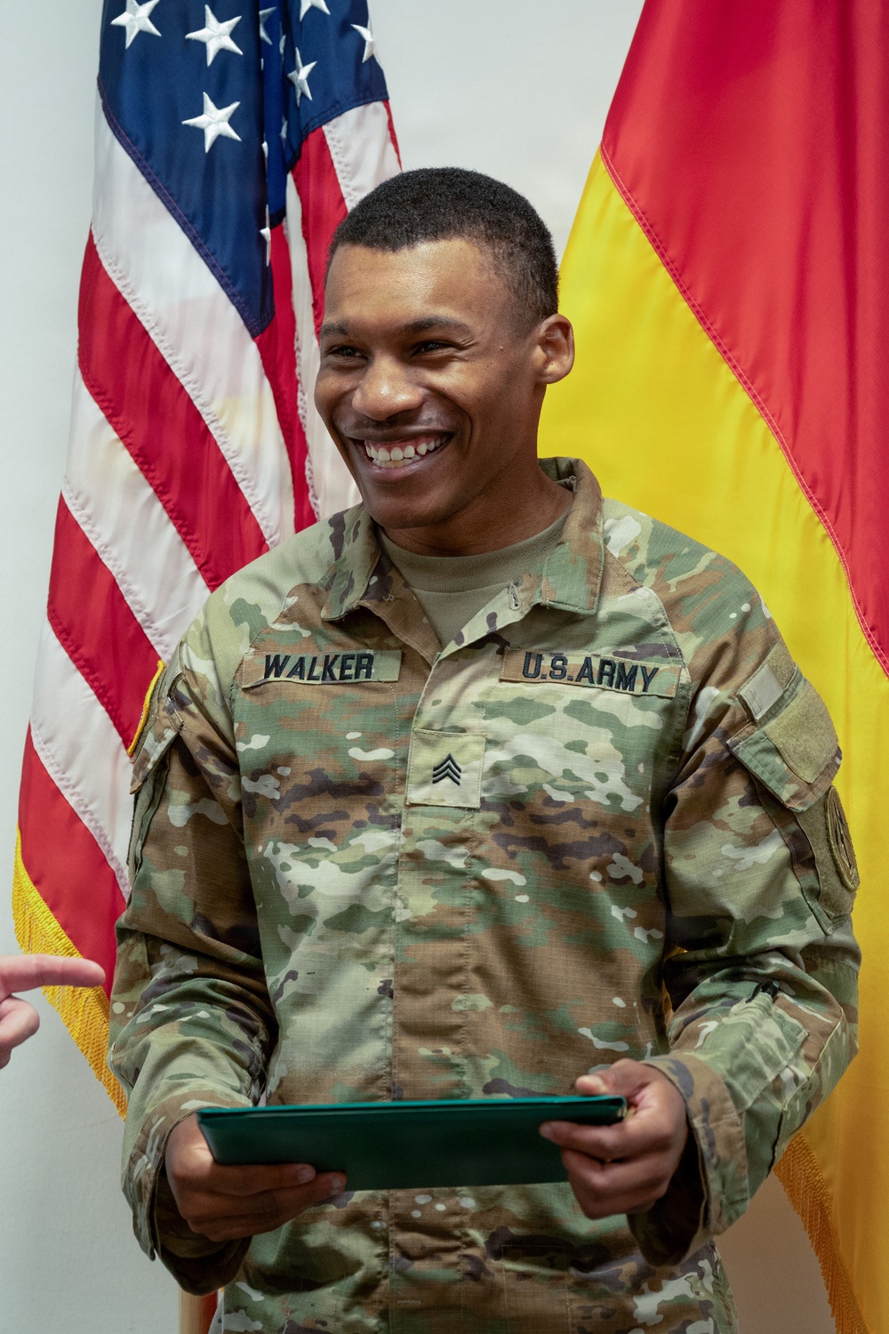 A U.S. Army Sergeant named Walker is smiling broadly while holding a green folder or certificate binder, standing in front of an American flag and what appears to be the German flag. He is wearing the Army Combat Uniform (ACU) in OCP (Operational Camouflage Pattern) with a Sergeant rank insignia on his chest. The setting appears to be a formal military ceremony or award presentation.