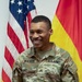 A U.S. Army Sergeant named Walker is smiling broadly while holding a green folder or certificate binder, standing in front of an American flag and what appears to be the German flag. He is wearing the Army Combat Uniform (ACU) in OCP (Operational Camouflage Pattern) with a Sergeant rank insignia on his chest. The setting appears to be a formal military ceremony or award presentation.