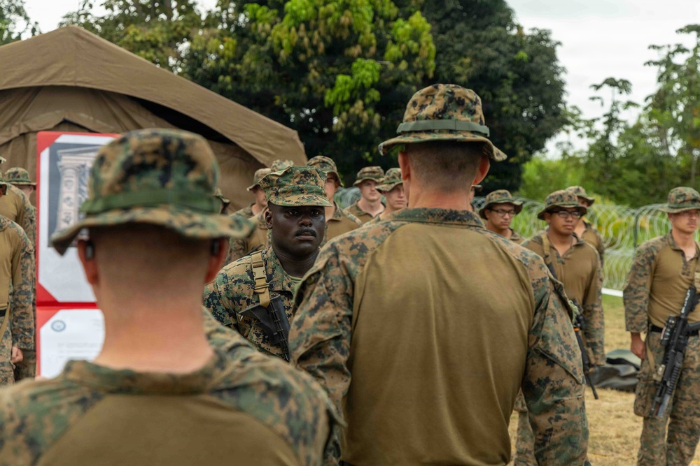 A group of military personnel in MARPAT (Marine pattern) camouflage uniforms are gathered in an outdoor setting near a military tent. A senior figure wearing a boonie hat appears to be addressing or briefing the assembled Marines. The setting suggests a field exercise or operational briefing at a forward operating base or training area.