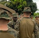 A group of military personnel in MARPAT (Marine pattern) camouflage uniforms are gathered in an outdoor setting near a military tent. A senior figure wearing a boonie hat appears to be addressing or briefing the assembled Marines. The setting suggests a field exercise or operational briefing at a forward operating base or training area.