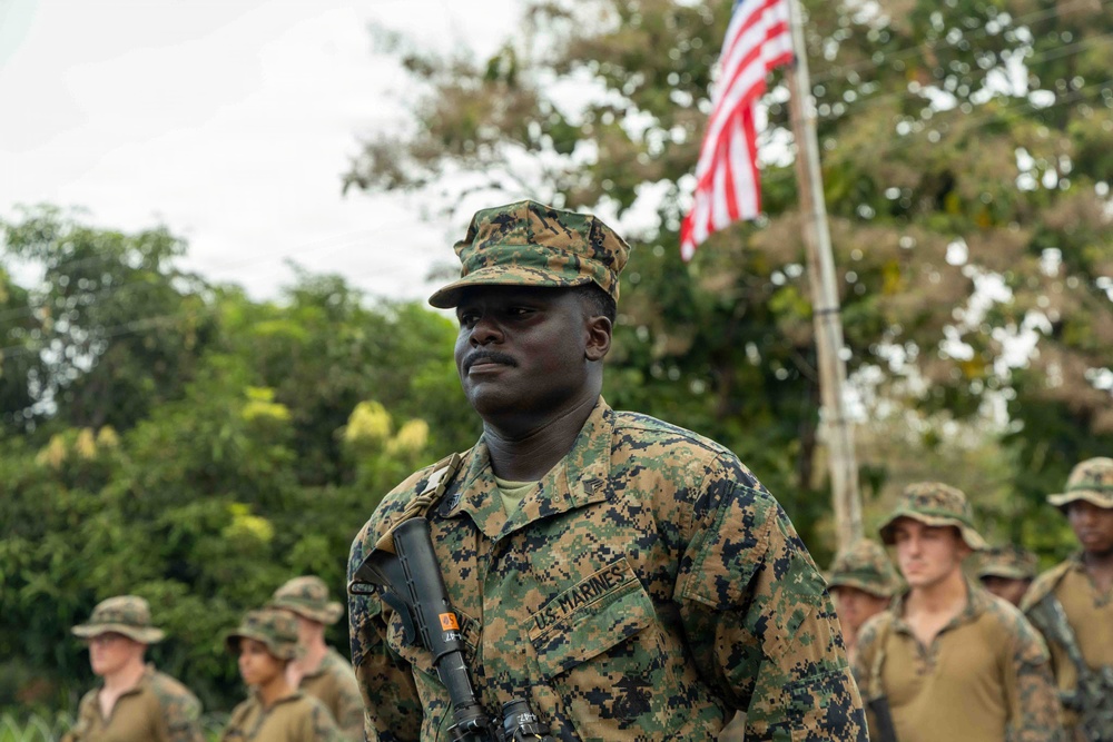 A U.S. Marine in the foreground stands at attention wearing MARPAT woodland camouflage uniform with a 'U.S. MARINES' chest tape and a patrol cap. An American flag is visible on a flagpole in the background, surrounded by tropical vegetation. Several other Marines in similar uniforms are visible in the background, suggesting a formation or ceremony.
