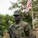 A U.S. Marine in the foreground stands at attention wearing MARPAT woodland camouflage uniform with a 'U.S. MARINES' chest tape and a patrol cap. An American flag is visible on a flagpole in the background, surrounded by tropical vegetation. Several other Marines in similar uniforms are visible in the background, suggesting a formation or ceremony.