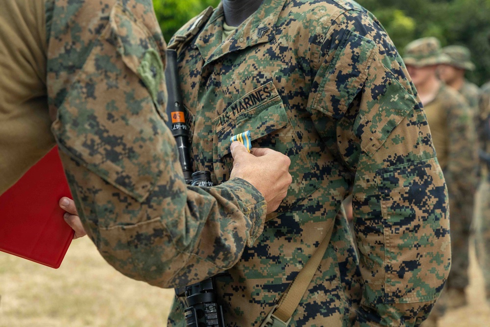 A U.S. Marines service member is receiving a military award or decoration during what appears to be an outdoor ceremony. A hand is pinning a ribbon or medal onto the chest of the Marine's MARPAT (Marine Pattern) digital camouflage uniform. Additional uniformed personnel are visible in the blurred background.