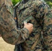 A U.S. Marines service member is receiving a military award or decoration during what appears to be an outdoor ceremony. A hand is pinning a ribbon or medal onto the chest of the Marine's MARPAT (Marine Pattern) digital camouflage uniform. Additional uniformed personnel are visible in the blurred background.