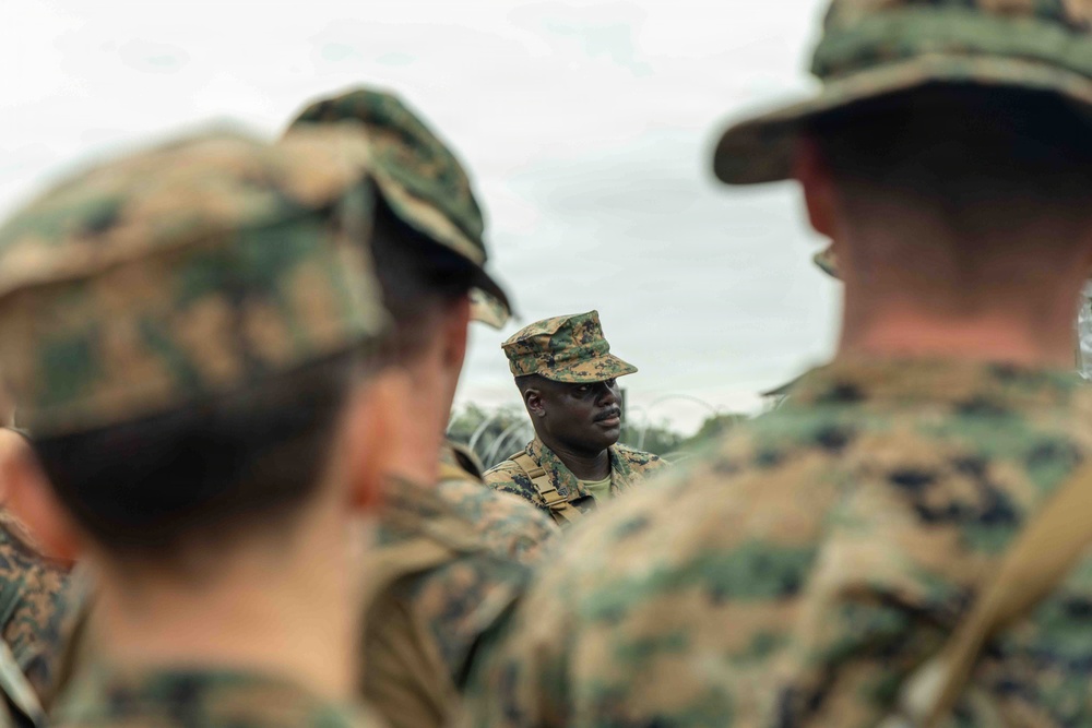 A group of military personnel in camouflage uniforms are gathered in what appears to be a briefing or instruction session outdoors. One individual in the center, in sharp focus, appears to be addressing or leading the group. The foreground figures are intentionally blurred, drawing attention to the central speaker.