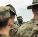 A group of military personnel in camouflage uniforms are gathered in what appears to be a briefing or instruction session outdoors. One individual in the center, in sharp focus, appears to be addressing or leading the group. The foreground figures are intentionally blurred, drawing attention to the central speaker.