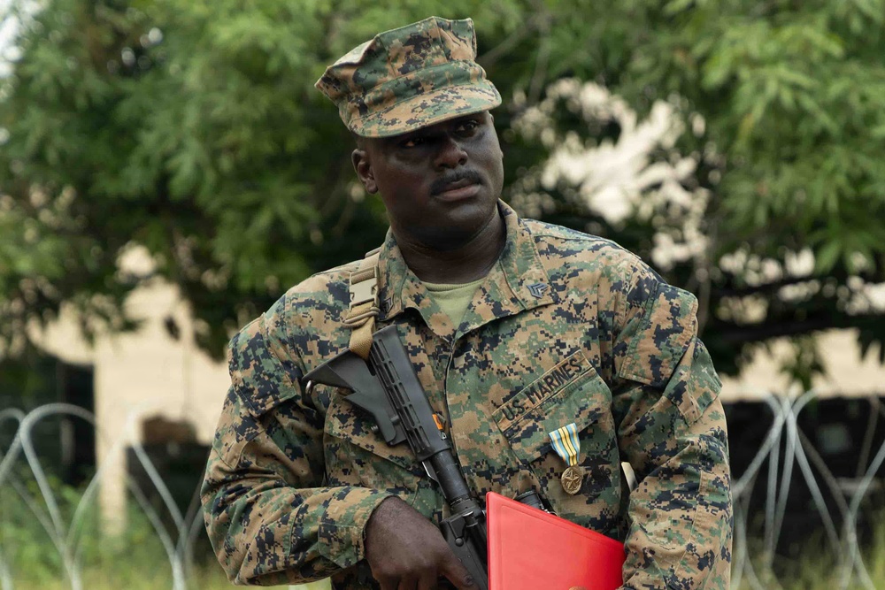 A U.S. Marine in MARPAT (Marine Pattern) digital camouflage uniform stands at attention outdoors. He is wearing a cover (cap), carrying a rifle slung across his body, and displaying a service medal on his chest. He holds a red folder/binder and appears to be in a formal or ceremonial setting.