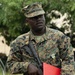 A U.S. Marine in MARPAT (Marine Pattern) digital camouflage uniform stands at attention outdoors. He is wearing a cover (cap), carrying a rifle slung across his body, and displaying a service medal on his chest. He holds a red folder/binder and appears to be in a formal or ceremonial setting.