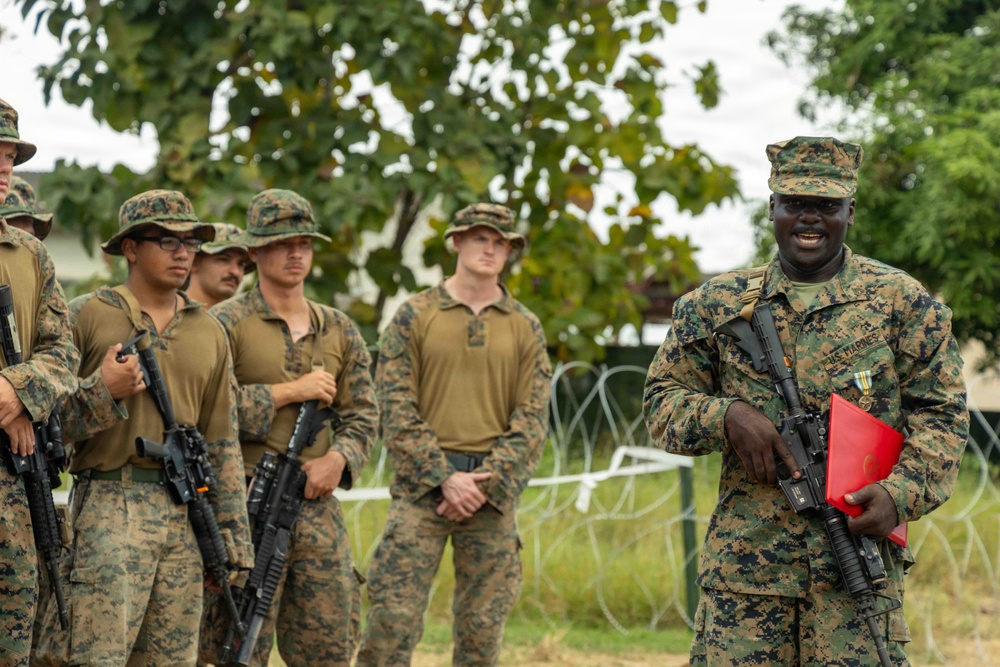 A group of U.S. Marines in MARPAT camouflage uniforms are assembled outdoors, with one senior Marine in the foreground appearing to address the group. The Marine in front is holding a red folder and has a rifle slung, and is wearing service ribbons on his uniform. The setting appears to be a field environment with concertina wire visible in the background.
