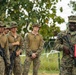 A group of U.S. Marines in MARPAT camouflage uniforms are assembled outdoors, with one senior Marine in the foreground appearing to address the group. The Marine in front is holding a red folder and has a rifle slung, and is wearing service ribbons on his uniform. The setting appears to be a field environment with concertina wire visible in the background.