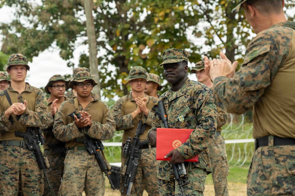 A U.S. Marine is being recognized in an outdoor military ceremony, holding a red presentation folder with the Marine Corps emblem and wearing a medal on his uniform. Fellow Marines in MARPAT camouflage uniforms are applauding him, while a senior Marine officer faces him from the right. The setting appears to be an outdoor field environment with trees in the background.