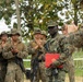 A U.S. Marine is being recognized in an outdoor military ceremony, holding a red presentation folder with the Marine Corps emblem and wearing a medal on his uniform. Fellow Marines in MARPAT camouflage uniforms are applauding him, while a senior Marine officer faces him from the right. The setting appears to be an outdoor field environment with trees in the background.