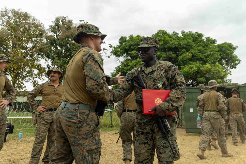Two U.S. Marines in MARPAT camouflage uniforms are shaking hands in what appears to be a field environment or forward operating base. The Marine on the right is holding a red folder bearing the U.S. Marines emblem and has a medal pinned to his chest, suggesting an award or recognition ceremony has taken place. Several other Marines in various states of uniform are visible in the background.