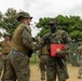 Two U.S. Marines in MARPAT camouflage uniforms are shaking hands in what appears to be a field environment or forward operating base. The Marine on the right is holding a red folder bearing the U.S. Marines emblem and has a medal pinned to his chest, suggesting an award or recognition ceremony has taken place. Several other Marines in various states of uniform are visible in the background.