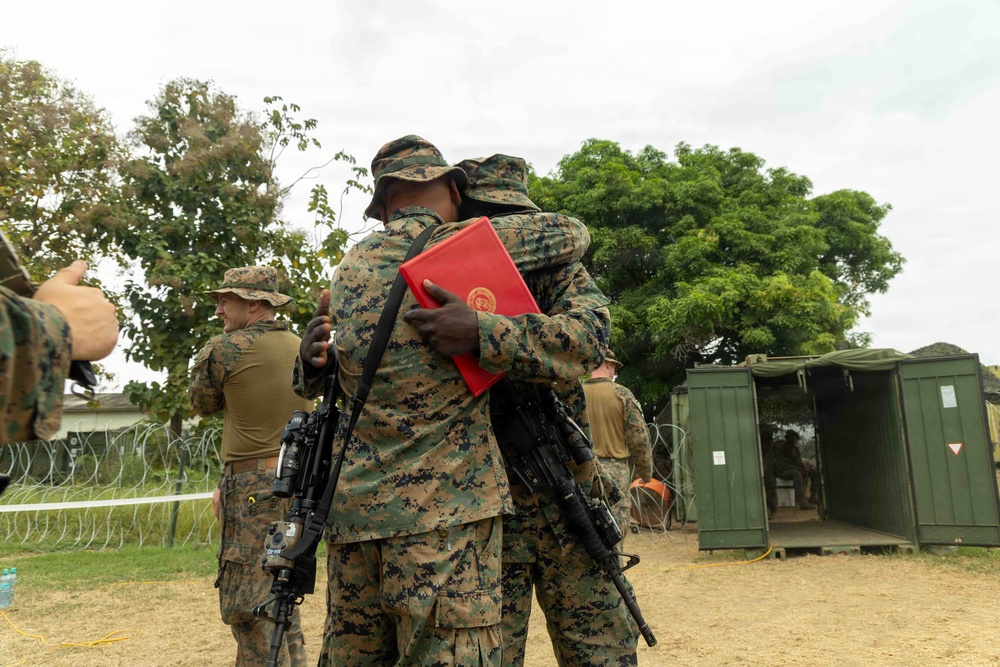 Two military personnel in MARPAT (Marine digital camouflage) uniforms are embracing in what appears to be a congratulatory or farewell hug at an outdoor military installation. One individual is holding a red folder or certificate, suggesting a formal recognition or promotion ceremony. The setting includes concertina wire perimeter fencing, military containers, and other uniformed personnel in the background.