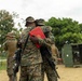 Two military personnel in MARPAT (Marine digital camouflage) uniforms are embracing in what appears to be a congratulatory or farewell hug at an outdoor military installation. One individual is holding a red folder or certificate, suggesting a formal recognition or promotion ceremony. The setting includes concertina wire perimeter fencing, military containers, and other uniformed personnel in the background.