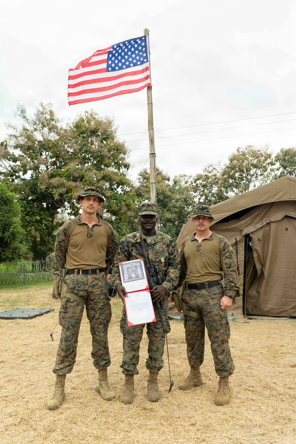 Three military personnel in U.S. Marine Corps MARPAT digital camouflage uniforms are posed in front of a raised American flag at what appears to be a forward operating base or field camp. The center individual is holding a red-covered certificate or award document and is armed with a rifle, suggesting this is a formal recognition or award ceremony. A military tent and concertina wire perimeter are visible in the background.