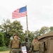 Three military personnel in U.S. Marine Corps MARPAT digital camouflage uniforms are posed in front of a raised American flag at what appears to be a forward operating base or field camp. The center individual is holding a red-covered certificate or award document and is armed with a rifle, suggesting this is a formal recognition or award ceremony. A military tent and concertina wire perimeter are visible in the background.