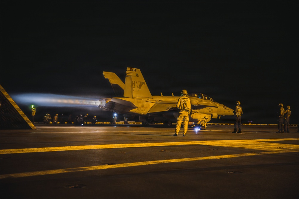 A U.S. Navy aircraft carrier flight deck at night showing a jet aircraft preparing for or undergoing catapult launch operations. The aircraft, numbered 504 with tail markings 'NF', has its engines at high thrust producing visible exhaust plumes. Multiple flight deck personnel in yellow jerseys are visible observing and managing the launch sequence.