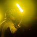 A person wearing a helmet with goggles and protective gear is holding two illuminated light wands in a dark, yellow-hued environment. The scene appears to depict a flight deck or airfield operations worker, likely a aircraft handler or director, using marshalling wands during nighttime operations. The yellow atmospheric glow and glowing wands create a dramatic, high-contrast visual.